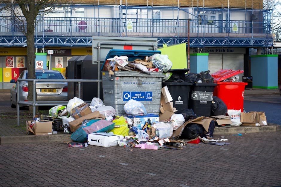 A collection of overflowing rubbish bins and scattered waste bags and packaging on a paved outdoor area in front of a commercial building. The scene includes a large grey mixed paper and cardboard recycling container with its lid partially open, revealing crumpled paper, newspapers, and cardboard boxes. Surrounding it are black and red waste bags, flattened cardboard boxes, and loose paper materials spilling onto the ground. There is a parked grey car nearby, partially obscured, and a metal railing separating the waste from the parked vehicle facing a busy retail strip with storefronts and signage in the background. The environment appears to be an urban or suburban parking area with minimal greenery, including a small tree to the left. Bright daylight illuminates the scene, highlighting the cluttered and unmanaged appearance of the waste, exemplifying an alternative waste handling situation that could benefit from professional rubbish removal services like those provided by Rubbish Removal Colliers Wood.