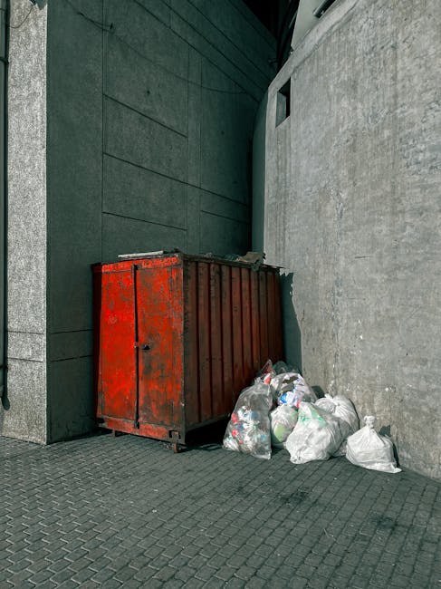 A large, rectangular metal skip with a weathered red finish, positioned against a grey, textured stone wall on a paved outdoor surface. The skip's surface shows signs of rust and paint chipping, indicating it's used for waste storage. Next to the skip, on the ground, there are multiple white plastic garbage bags filled with rubbish, some slightly crumpled and loosely tied. The scene is set in an urban or commercial area, with a dark green wall and a small, rectangular window visible in the background. The lighting appears natural, possibly overcast, with no shadows cast, and the setting suggests an environment suitable for waste collection or private rubbish removal services, such as those provided by Rubbish Removal Colliers Wood, facilitating alternative waste handling outside of standard public waste collection systems.