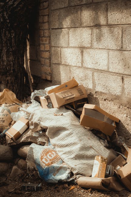 A collection of overflowing rubbish bins and scattered waste bags and packaging on a paved outdoor area in front of a commercial building. The scene includes a large grey mixed paper and cardboard recycling container with its lid partially open, revealing crumpled paper, newspapers, and cardboard boxes. Surrounding it are black and red waste bags, flattened cardboard boxes, and loose paper materials spilling onto the ground. There is a parked grey car nearby, partially obscured, and a metal railing separating the waste from the parked vehicle facing a busy retail strip with storefronts and signage in the background. The environment appears to be an urban or suburban parking area with minimal greenery, including a small tree to the left. Bright daylight illuminates the scene, highlighting the cluttered and unmanaged appearance of the waste, exemplifying an alternative waste handling situation that could benefit from professional rubbish removal services like those provided by Rubbish Removal Colliers Wood.