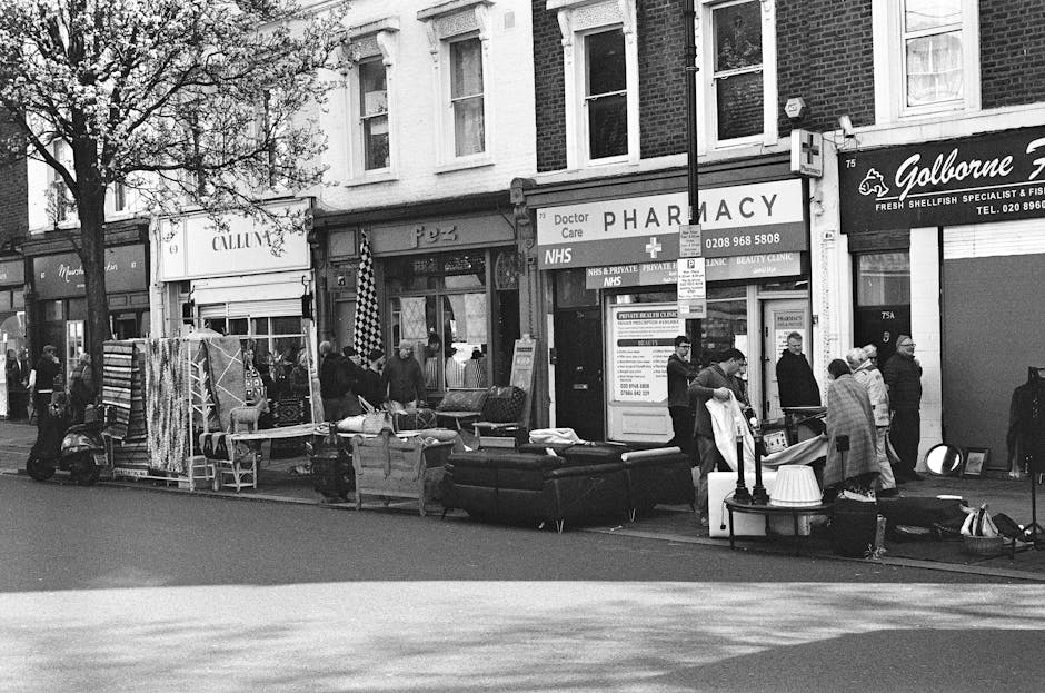 A busy street scene in front of a row of shops features an outdoor market setup with various secondhand furniture items and household goods arranged on the pavement, including a black sofa and a small table with a white lampshade. Several people are gathered around the display, some examining items, while others appear to be engaged in conversation or walking past. A few individuals are standing near a parked scooter. The background shows a pharmacy with signage indicating NHS services, a fish and seafood shop, and other commercial storefronts, all within a traditional brick building facaded with multiple windows. Trees line the street on the left side of the image, providing some canopy, and a lamp post extends into the scene. The environment appears to be during day time, with natural light casting soft shadows. The scene, captured in black and white, illustrates a typical marketplace or community street where items are being displayed for sale or clearance, aligning with the concept of private waste handling or alternative rubbish collection methods. Rubbish Removal Colliers Wood might be involved in clearing larger items or assisting outdoor clearance of such waste items.