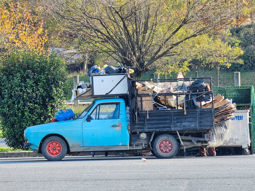 A small, light blue flatbed truck parked parallel to the curb on a street during daylight hours, loaded with various types of rubbish and waste materials. The truck's bed is filled with cardboard boxes, plastic bags, and other debris, some of which are secured with rope or tied to the sides. The vehicle's wheels are painted red, and it has a worn, slightly faded finish. Behind the truck, there is a large tree with spreading branches and greenish-yellow leaves, indicating a possible autumn setting. To the left of the truck, a dense green bush partially obscures the front wheel, while the background features a fenced area and additional foliage. The environment appears to be a residential or suburban area with natural surroundings. The scene suggests an example of private waste collection or rubbish removal, typical of services offered by companies like Rubbish Removal Colliers Wood.