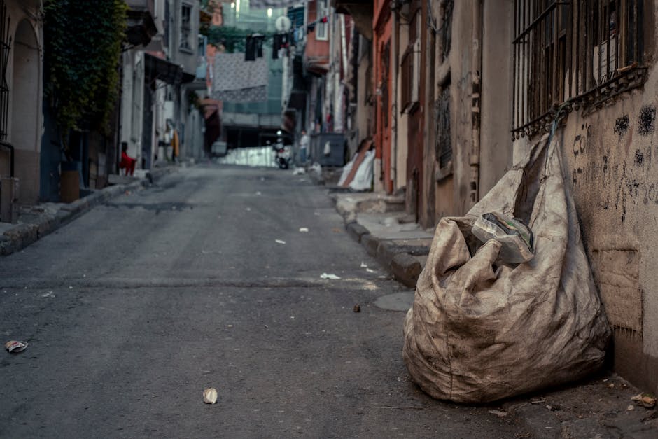 A large, dirty, beige cloth bag filled with mixed rubbish rests against a wall on a narrow, inclined urban street. The bag, made of coarse fabric, appears worn and stained, with a pile of discarded items visible at the top, including plastic packaging and overused gloves. The street surface is uneven with patches of asphalt and scattered debris, such as crumpled paper and small litter. On the left side, the pavement is bordered by a low curb, and on the right, there are metal bars attached to the weathered wall displaying graffiti. In the background, the street slopes upward, lined with tightly packed residential buildings of various materials and muted colours, some with hanging laundry and exposed brickwork. Overcast lighting creates a subdued atmosphere typical of areas where independent waste handling might be necessary, highlighting the presence of informal rubbish disposal. This scene exemplifies typical urban waste that Rubbish Removal Colliers Wood might manage through private rubbish collection services in a densely built environment.
