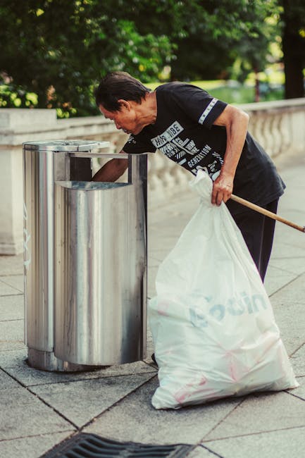 A man with dark hair, wearing a black T-shirt with white text, is bent over an outdoor stainless steel rubbish bin, actively depositing waste. He is holding a large white garbage bag with both hands, with the bag appearing to be partially filled. The scene takes place on a paved area, possibly in a park or public space, with lush green trees and foliage in the background. The rubbish bin is cylindrical with a metallic finish, situated next to a stone or concrete wall with a decorative railing. The environment is well-lit, suggesting daytime, and the scene reflects routine waste collection or disposal activity, aligning with the services of Rubbish Removal Colliers Wood in providing alternative waste handling solutions.
