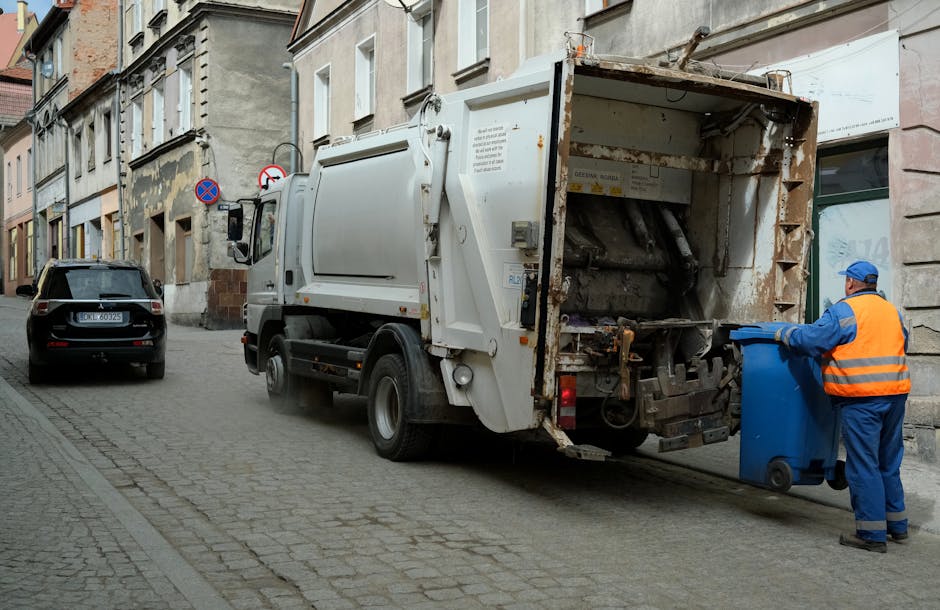 A white commercial rubbish collection truck is parked on a narrow urban street, with its rear loading compartment open, revealing mechanical components and a rusty interior. The truck is positioned next to a sidewalk with cobblestone paving, adjacent to old, multi-story buildings with a weathered appearance, some featuring peeling paint and exposed brickwork. To the right of the truck, a worker wearing a blue cap, orange high-visibility vest, and blue overalls is standing beside a blue waste bin, actively tipping or emptying contents into the truck's rear compartment. Behind the truck, a black car is parked parallel to the curb, and further along the street are additional parked vehicles. The scene captures a daytime setting with natural light, emphasizing the industrial and functional nature of on-site waste disposal, consistent with private rubbish removal services provided by companies such as Rubbish Removal Colliers Wood, operating in the area for alternative waste handling and local refuse collection.