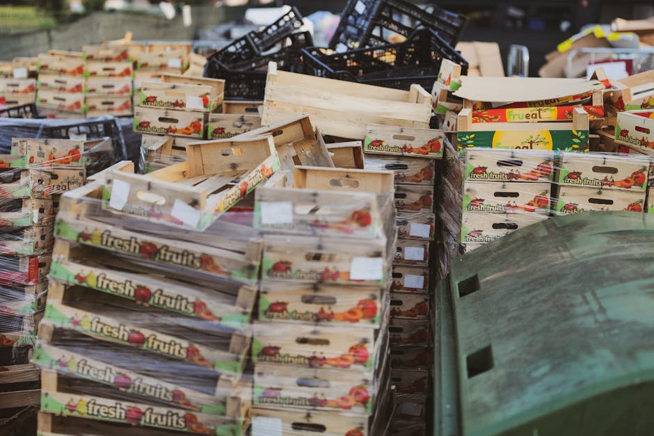 A large collection of empty cardboard fruit boxes, predominantly labeled with 'fresh fruits' and colorful images of produce, are stacked and arranged in an outdoor area. The boxes vary in condition, with some appearing slightly worn or unfolded, and are positioned in multiple directions, creating a cluttered scene typical of a waste management or rubbish removal operation. In the foreground, a green waste bin with a textured surface and small rectangular holes is visible, partially obscuring the lower right corner of the image. Behind the boxes, black plastic crates are stacked haphazardly, adding to the disorderly appearance of the scene. Natural daylight illuminates the scene, highlighting the textures of the cardboard and the slight weathering of the materials. The environment suggests an outdoor storage space or back area where waste or surplus stock is temporarily held before disposal or recycling, aligned with services offered by Rubbish Removal Colliers Wood specializing in alternative waste handling solutions.