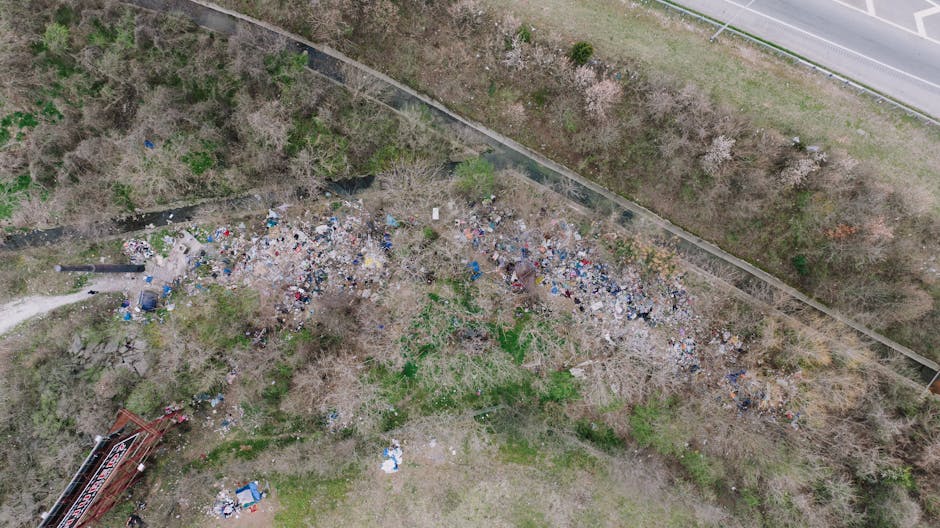 An aerial view of an outdoor area showing a large, uneven stretch of ground covered with scattered piles of mixed waste, including plastic bags, debris, and miscellaneous rubbish. The waste appears to be a combination of household and industrial materials, with some items partially buried or entangled among leafless trees and shrubbery. To the right, there is a paved road with a white line marking the edge, and a concrete wall runs parallel to the road, separating the rubbish-strewn area from the street. Adjacent to the waste, sparse greenery and patches of grass are visible, with the ground showing signs of natural wear and debris accumulation. A red construction or waste container is located toward the lower left corner, with some smaller scattered items nearby. The scene suggests an unauthorised dumping site or neglected unmanaged waste. For private rubbish removal services, companies such as Rubbish Removal Colliers Wood may assist with clearing such outdoor areas, especially where access issues limit conventional collection. The natural environmental surroundings and the proximity to the road highlight the importance of controlled waste management through independent collection or on-site clearance solutions.