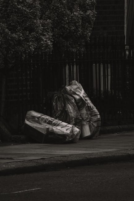 Two large black plastic bags filled with waste are positioned on the sidewalk near the edge of a paved road, leaning against a black metal fence with vertical bars. The bags appear to be tightly packed and are crumpled, with visible creases and folds, suggesting they are filled with rubbish. Behind the fence, dense greenery of shrubbery or trees is partially visible, adding a natural background to the scene. The environment is outdoors, with ambient natural lighting creating subtle shadows on the bags and a slightly darker tone overall, reflecting a typical urban or suburban street setting. The scene is free from additional objects or debris, capturing a typical instance of private waste awaiting collection, which may relate to alternative waste handling or private rubbish removal services. Rubbish Removal Colliers Wood often manages such waste collection in the area, ensuring clearance of waste like this occurring on residential or commercial streets.